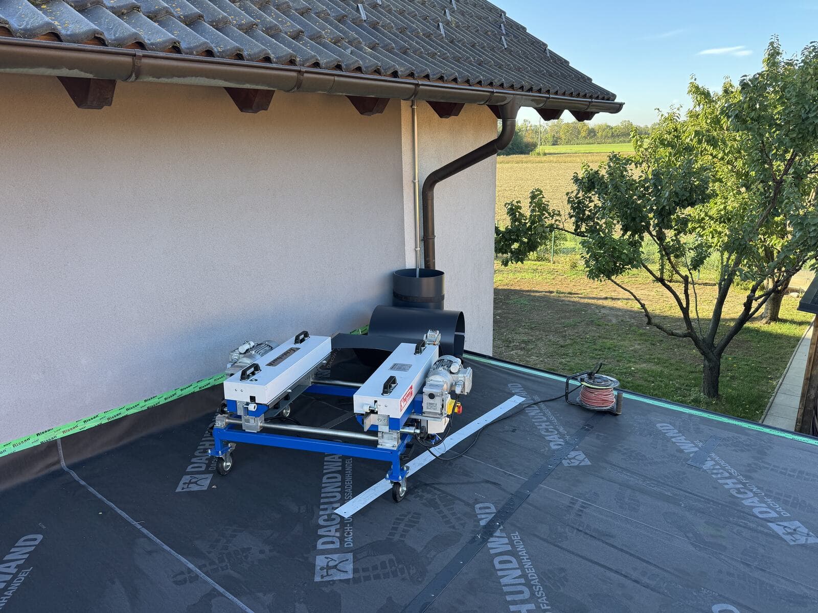 Die mobile Profilieranlage auf ihrem blauen Rollgestell steht auf der vorbereiteten Dachfläche neben dem Wohnhaus.